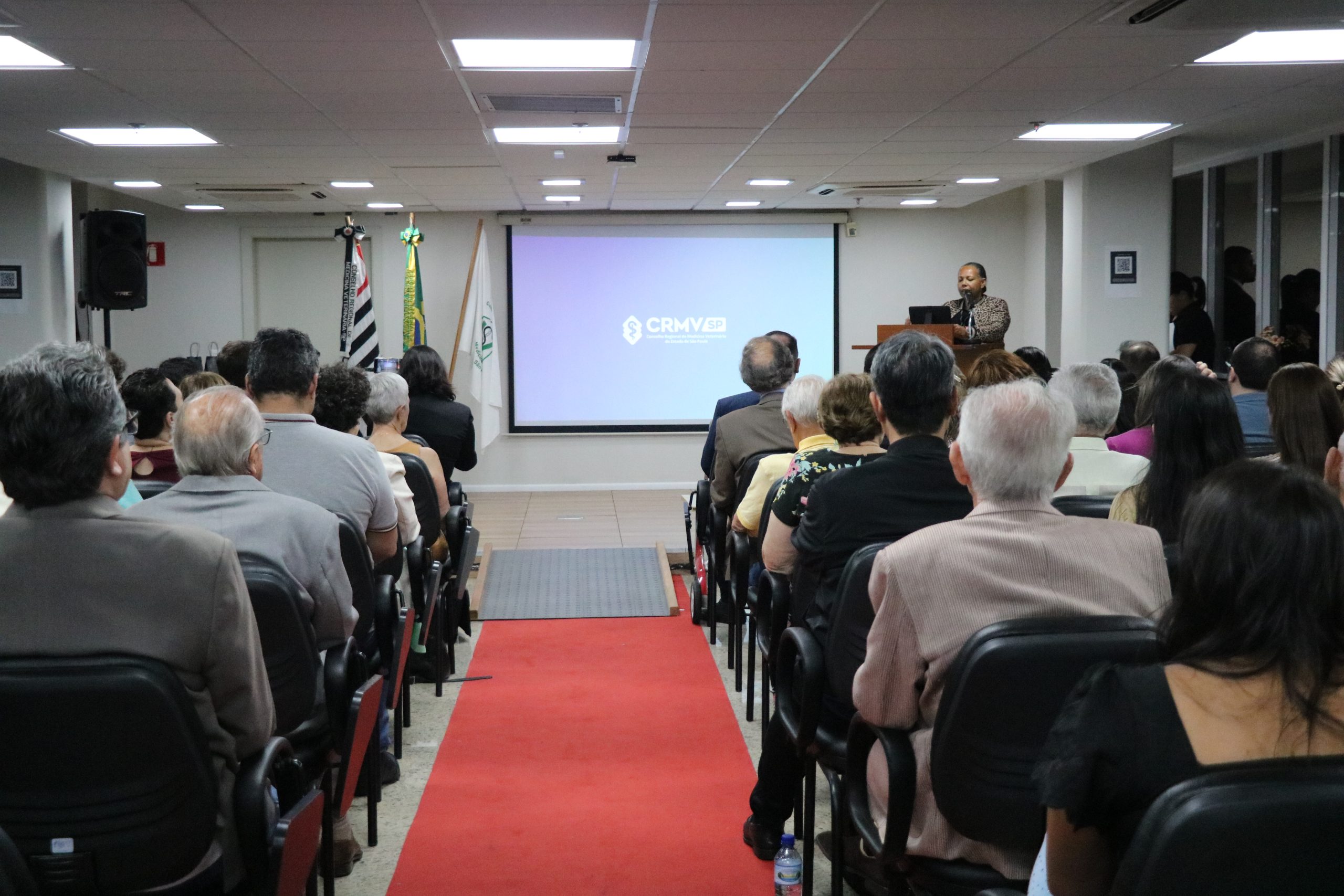 No auditório da sede, homens e mulheres assistem à apresentação de conselheira e presidente da Comissão de Avaliação e Julgamento que deu início à cerimônia de premiação.