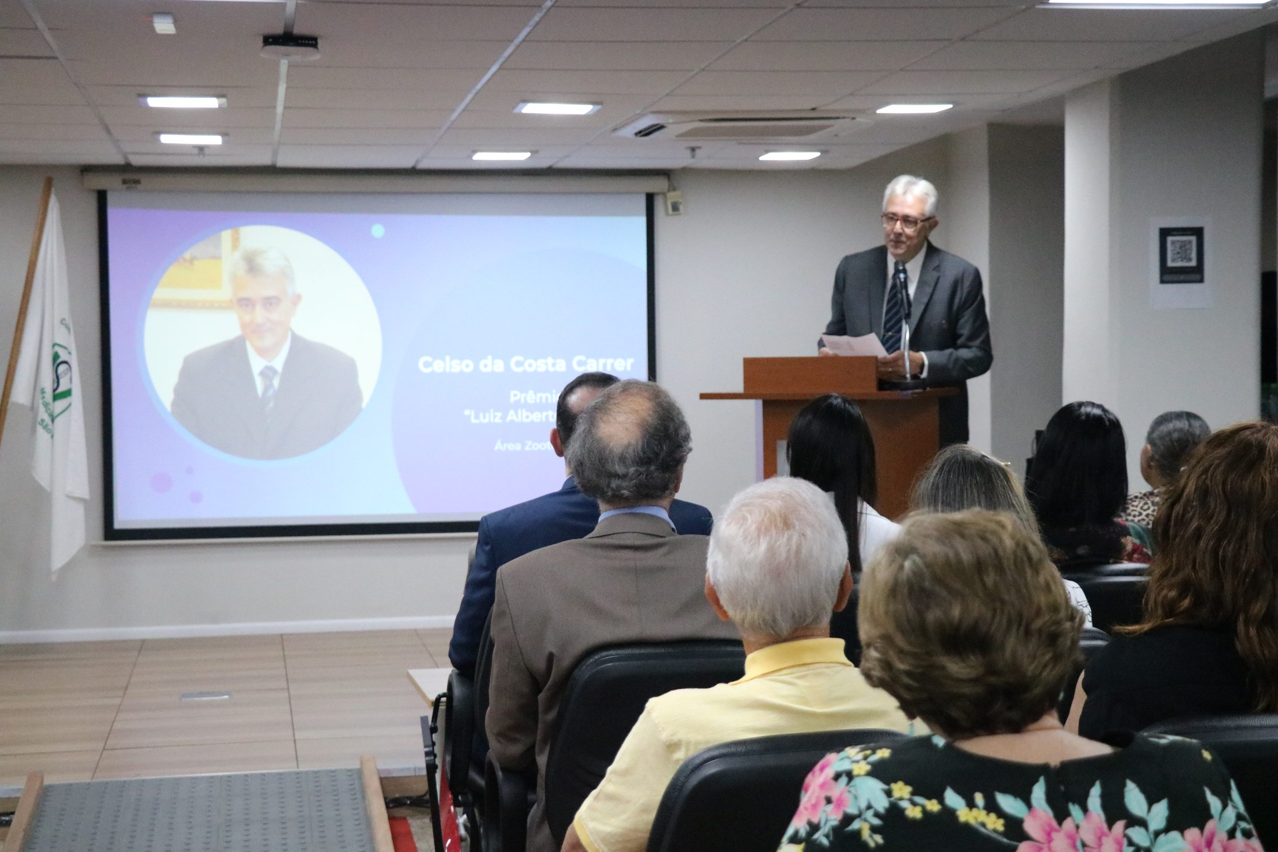 O premiado na categoria Zootecnia discursa aos presentes no auditório, ele está no púlpito e ao fundo sua foto aparece no telão.