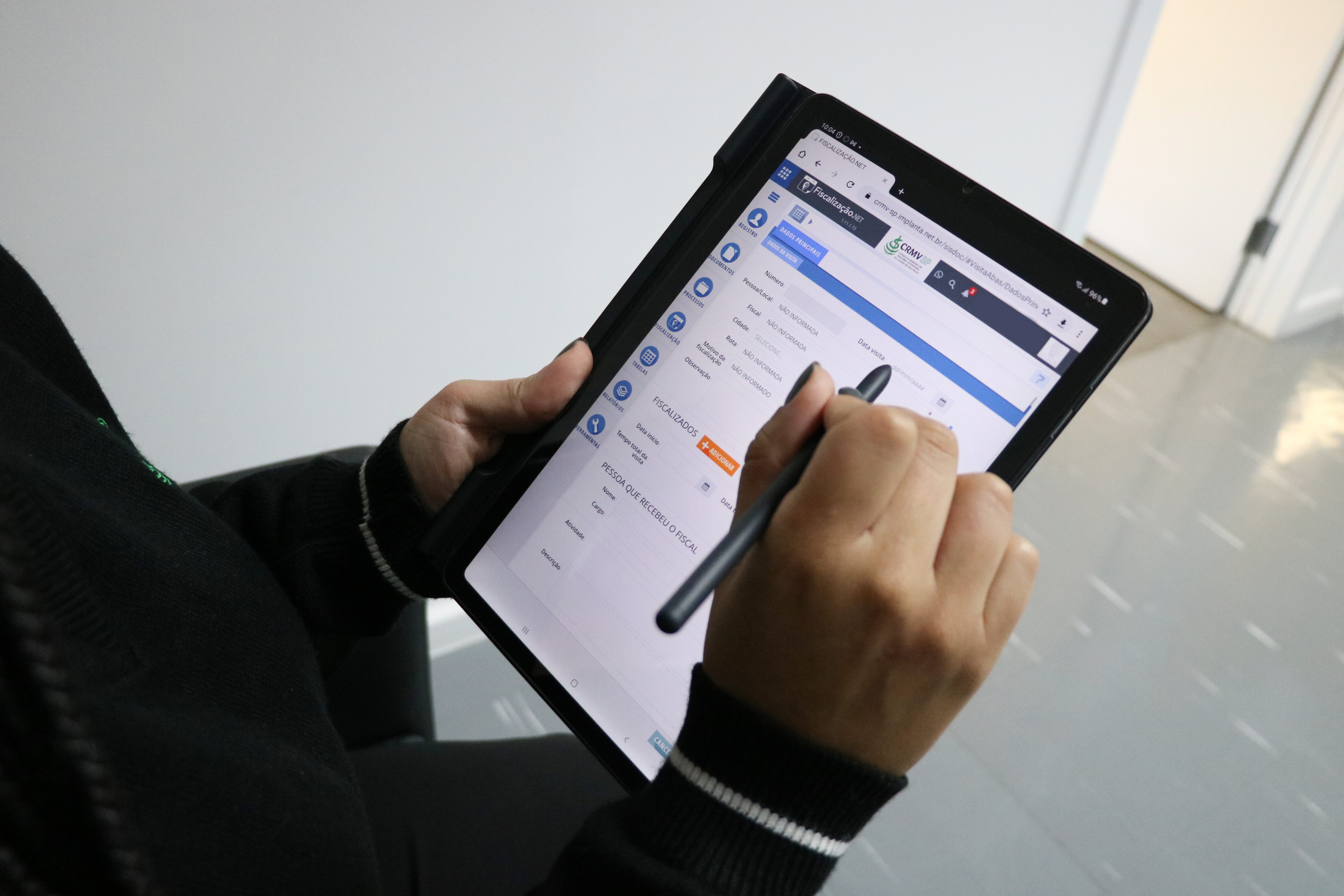 Mãos femininas segurando caneta digital e tablet preto que está acessando sistema de fiscalização do CRMV-SP.