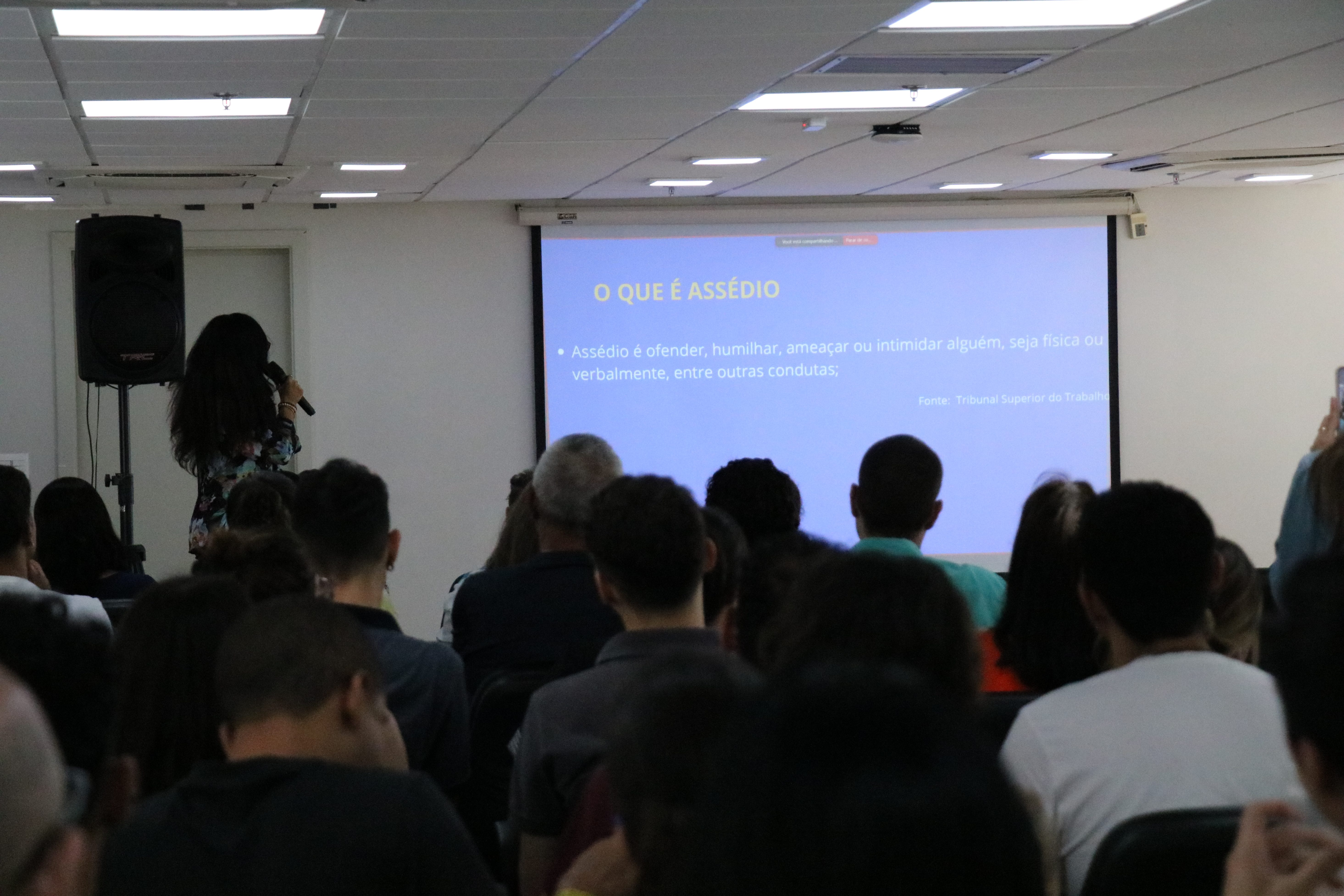 Pessoas assistindo uma mulher falando em frente a tela.