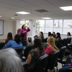 Na foto há uma mulher de camiseta e calça rosa, óculos, cabelos pretos, ondulados e de pele branca. ela está falando ao microfone junto ao público.