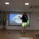 Na foto há uma mulher de camiseta preta, cabelos longos e ondulados, pele branca, óculos e calça bege falando ao microfone no palco.