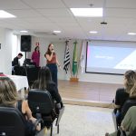 Na foto há uma mulher de camiseta marrom e calça azul escura falando ao microfone encima do palco para o público.