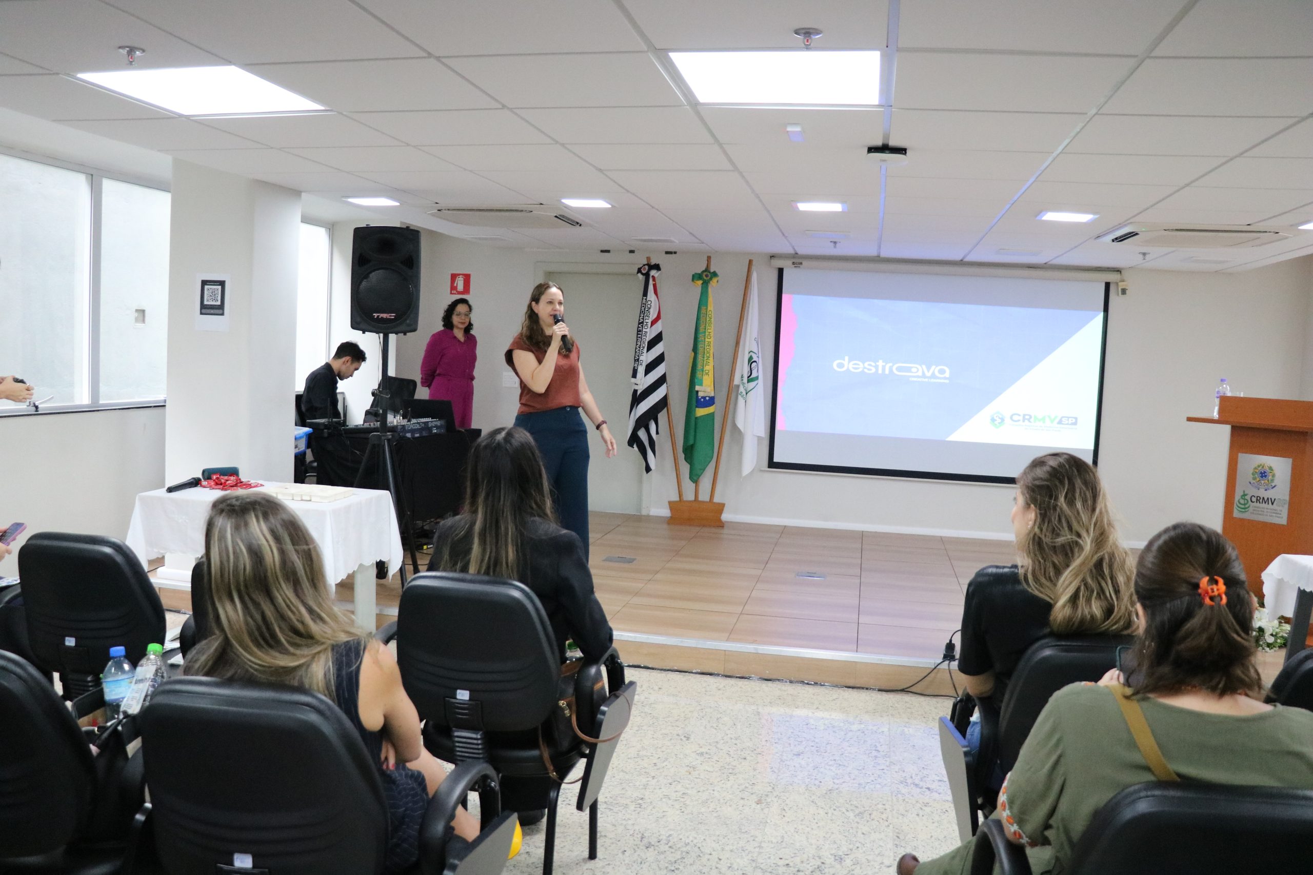 Na foto há uma mulher de camiseta marrom e calça azul escura falando ao microfone encima do palco para o público.