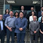Doze pessoas em pé na escadaria em frente a um prédio comercial cinza em Belo Horizonte. Entre eles, a vice presidente Carolina Saraiva Filippos de Toledo, que veste calça, blusa e blazer pretos, o diretor técnico Leonardo Burlini Soares e o chefe jurídico, Bruno Fassoni.
