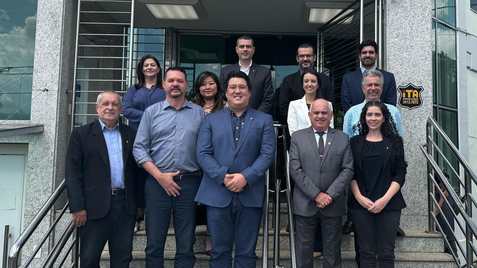 Doze pessoas em pé na escadaria em frente a um prédio comercial cinza em Belo Horizonte. Entre eles, a vice presidente Carolina Saraiva Filippos de Toledo, que veste calça, blusa e blazer pretos, o diretor técnico Leonardo Burlini Soares e o chefe jurídico, Bruno Fassoni.
