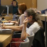 Mulher de cabelos loires de óculos sentada, frente ao computador.