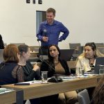 Três mulheres sentadas conversando e um homem de pé com cabelo curto e camisa azul segurando microfone.