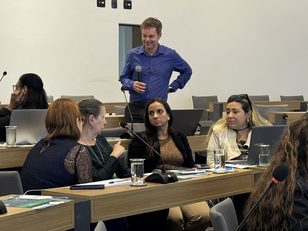 Três mulheres sentadas conversando e um homem de pé com cabelo curto e camisa azul segurando microfone.