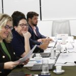 Duas mulheres e um homem sentados a uma mesa com computador para cada.