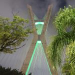 Vista lateral da ponte estaiada iluminada de verde em homenagem ao dia do zootecnista. Aparece à esquerda e à direita as copas de duas árvores.