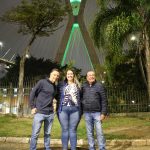 Os conselheiros: Martin Jacques Cavaliero, que veste calça jeans azul e casaco de moleton azul marinho, Daniela Denadai, que veste calça jeans azul, blusa branca com estampa de folhas azuis e casaco azul marinho e Haroldo Alberti que veste calça jeans e casaco preto, em pé sorridentes pousando para foto em frente à ponte estaiada iluminada de verde em homenagem ao dia do zootecnista.