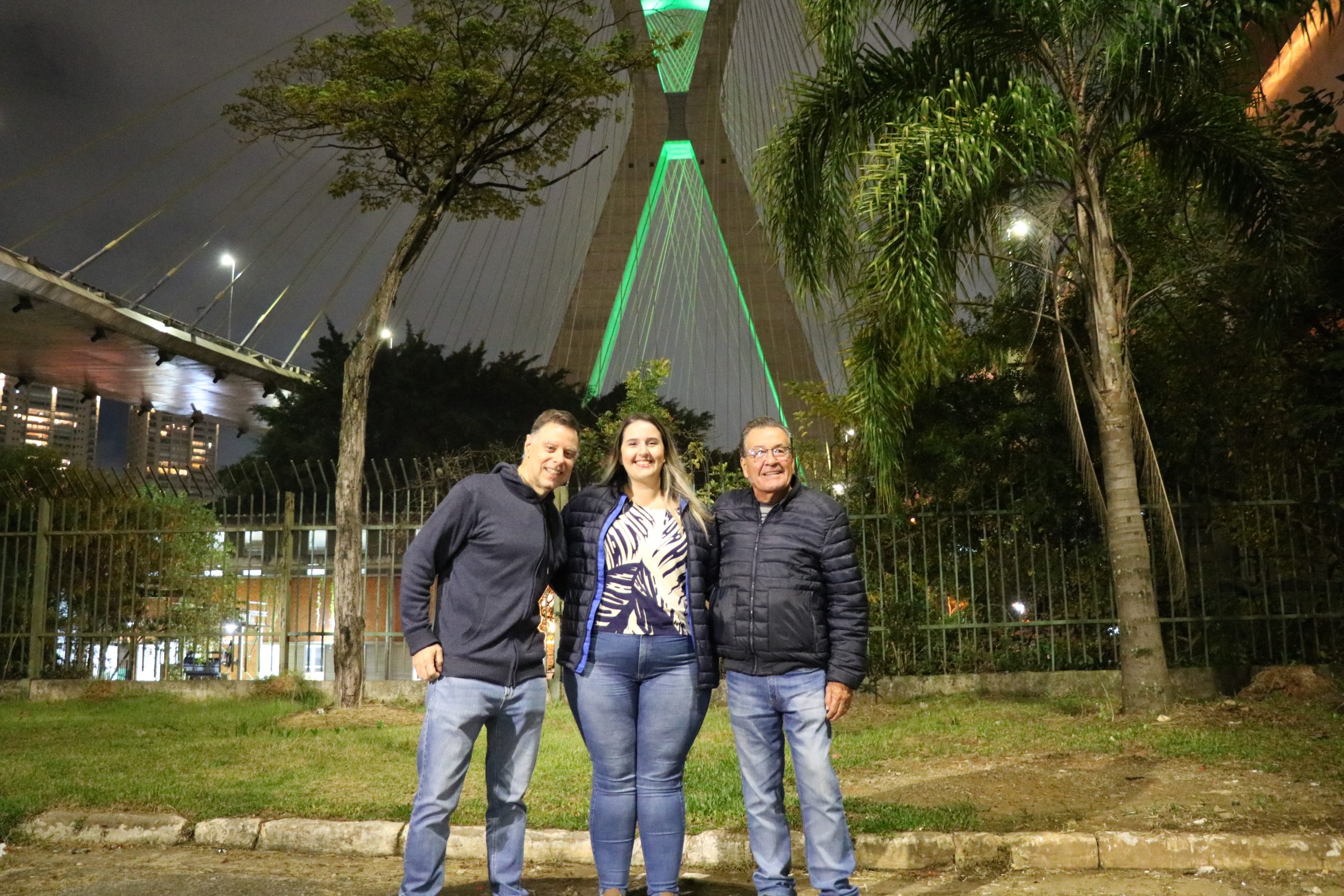 Os conselheiros: Martin Jacques Cavaliero, que veste calça jeans azul e casaco de moleton azul marinho, Daniela Denadai, que veste calça jeans azul, blusa branca com estampa de folhas azuis e casaco azul marinho e Haroldo Alberti que veste calça jeans e casaco preto, em pé sorridentes pousando para foto em frente à ponte estaiada iluminada de verde em homenagem ao dia do zootecnista.