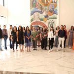 Homens e mulheres reunidos para foto. Ao fundo, uma obra de arte feita em azulejos.