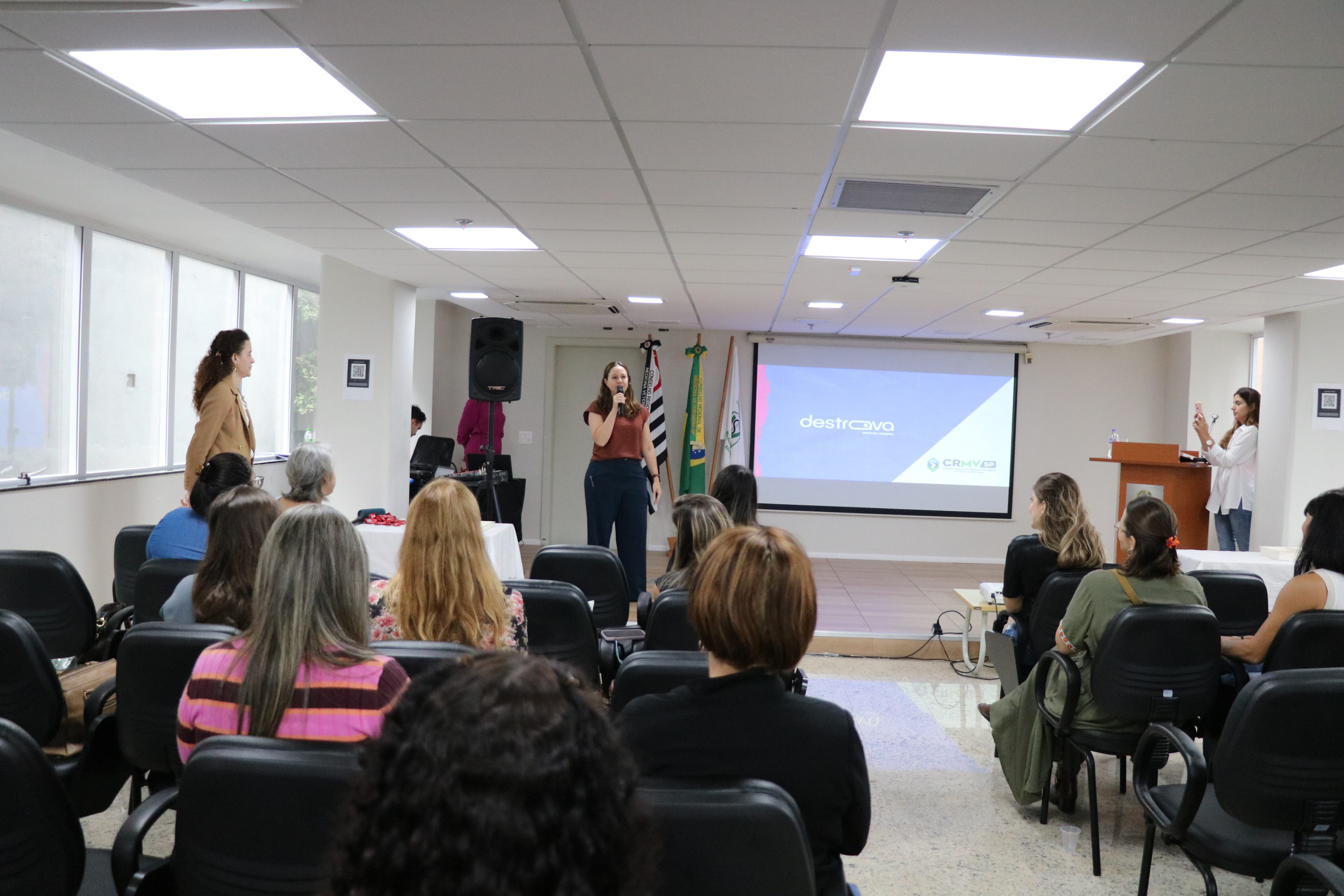 Na foto há uma mulher de blusa marrom e calça azul escura falando ao microfone encima do palco para o público.
