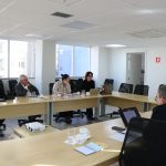 Sala de reunião com pessoas sentadas conversando. Em cima da mesa, cadernos, canetas, computadores, xícaras de café e copos de vidro com água. Ao fundo, uma bandeira do Brasil e outra do estado de São Paulo.