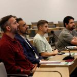 Pessoas sentadas a mesa assistindo a uma apresentação.