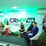 Pessoas em um palco durante cerimônia de posse do CRMV Maranhão. Da esquerda para a direita, três bandeiras, homem pardo sentado em uma cadeira branca, vestindo terno blusa e sapatos pretos; mulher loira branca de fios loiros longos usando vestido preto e dourado, óculos de lente transparente e sapato de bico fino branco; ao lado, mulher parda de fios pretos lisos e volumosos na altura dos ombros vestindo calça vermelha, blusa branca e blazer creme, ela usa óculos preto, ao lado uma cadeira branca vazia, ao lado, mulher branca de fios loiros claros ondulados, ela veste vestido preto justo na altura do joelho e sandália branca, ao lado de homem branco de barba longa grisalha, vestindo calça jeans escura, blusa preta e bota marrom, do lado do atual presidente do CRMV Maranhão, que é um homem de fios pretos, cabelo claro e veste terno azul, blusa branca e sapato preto. Em pé, um homem calvo de terno cinza, blusa branca e gravata verde fala ao microfone.