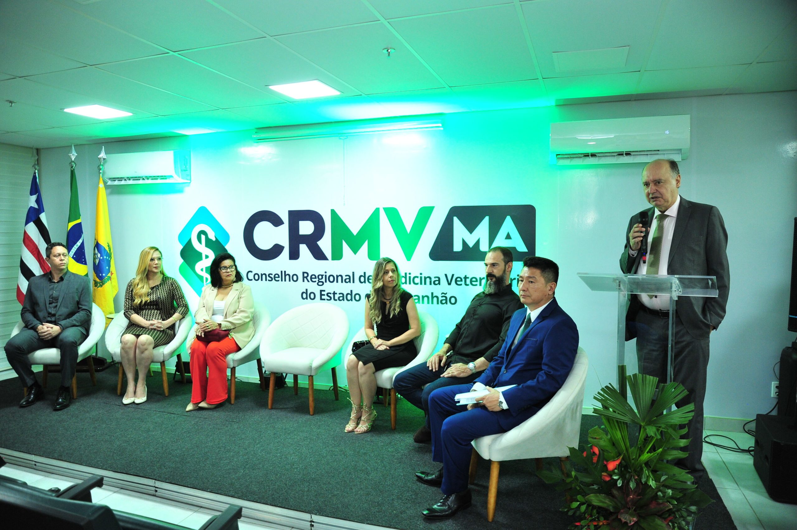 Pessoas em um palco durante cerimônia de posse do CRMV Maranhão. Da esquerda para a direita, três bandeiras, homem pardo sentado em uma cadeira branca, vestindo terno blusa e sapatos pretos; mulher loira branca de fios loiros longos usando vestido preto e dourado, óculos de lente transparente e sapato de bico fino branco; ao lado, mulher parda de fios pretos lisos e volumosos na altura dos ombros vestindo calça vermelha, blusa branca e blazer creme, ela usa óculos preto, ao lado uma cadeira branca vazia, ao lado, mulher branca de fios loiros claros ondulados, ela veste vestido preto justo na altura do joelho e sandália branca, ao lado de homem branco de barba longa grisalha, vestindo calça jeans escura, blusa preta e bota marrom, do lado do atual presidente do CRMV Maranhão, que é um homem de fios pretos, cabelo claro e veste terno azul, blusa branca e sapato preto. Em pé, um homem calvo de terno cinza, blusa branca e gravata verde fala ao microfone.