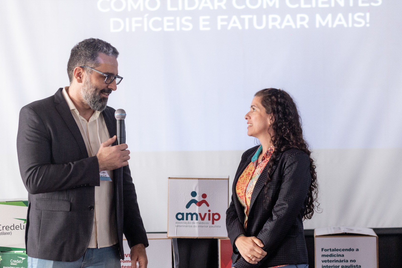 Homem em pé de blazer com microfone falando com a vice presidente do CRMV-SP, Carolina Filippos.