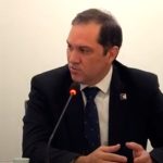 Homem de terno preto e cabelo curto sentado falando ao microfone.