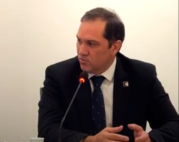 Homem de terno preto e cabelo curto sentado falando ao microfone.