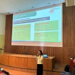 Mulher falando no microfone frente ao telão no palco. Ela veste calça preta e blazer creme com botões dourados. É a dra. Carla Maria Figueiredo.