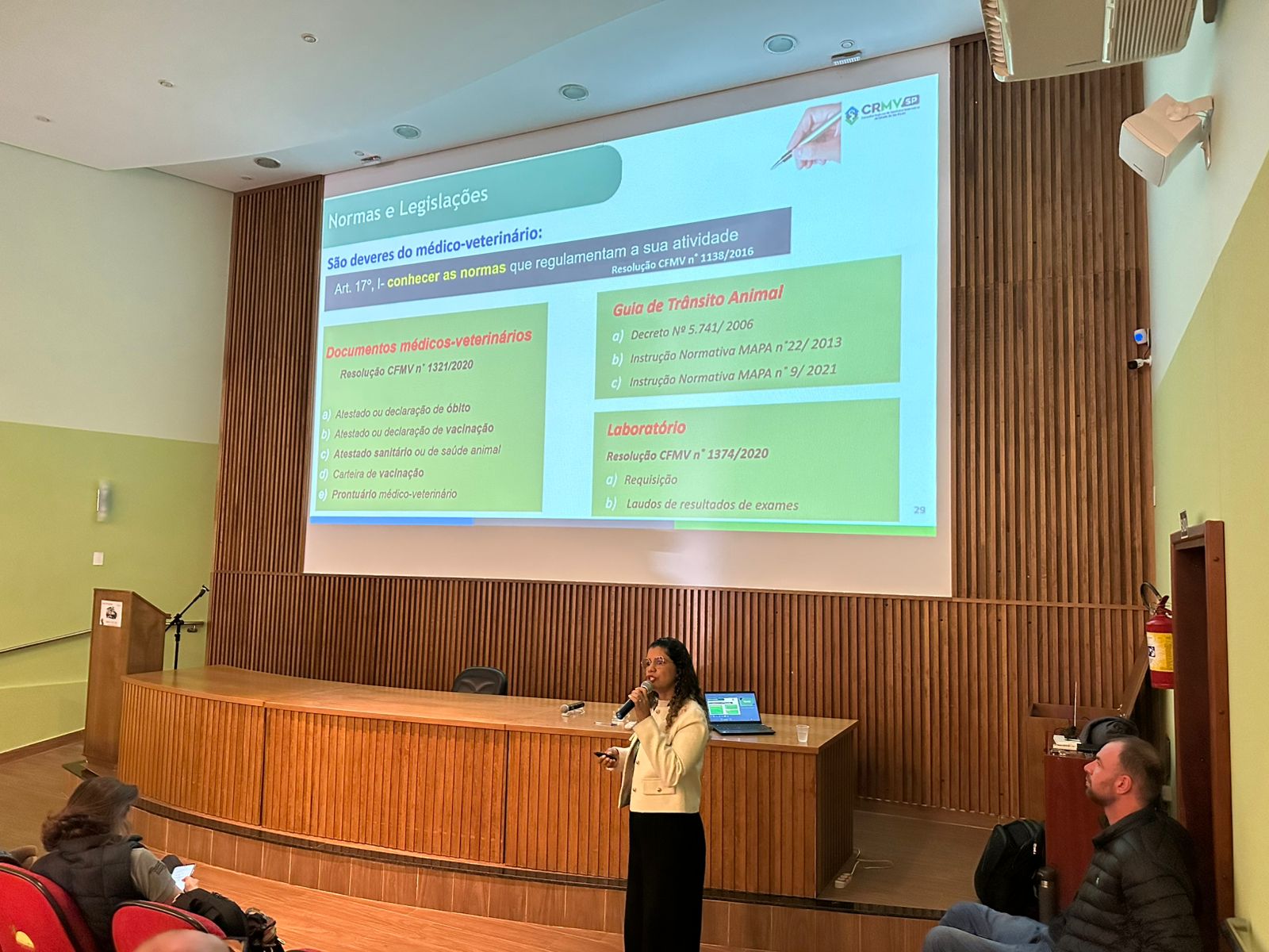 Mulher falando no microfone frente ao telão no palco. Ela veste calça preta e blazer creme com botões dourados. É a dra. Carla Maria Figueiredo.