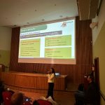 Mulher falando no microfone frente ao telão no palco. Ela veste calça preta e blazer creme com botões dourados. É a dra. Carla Maria Figueiredo.