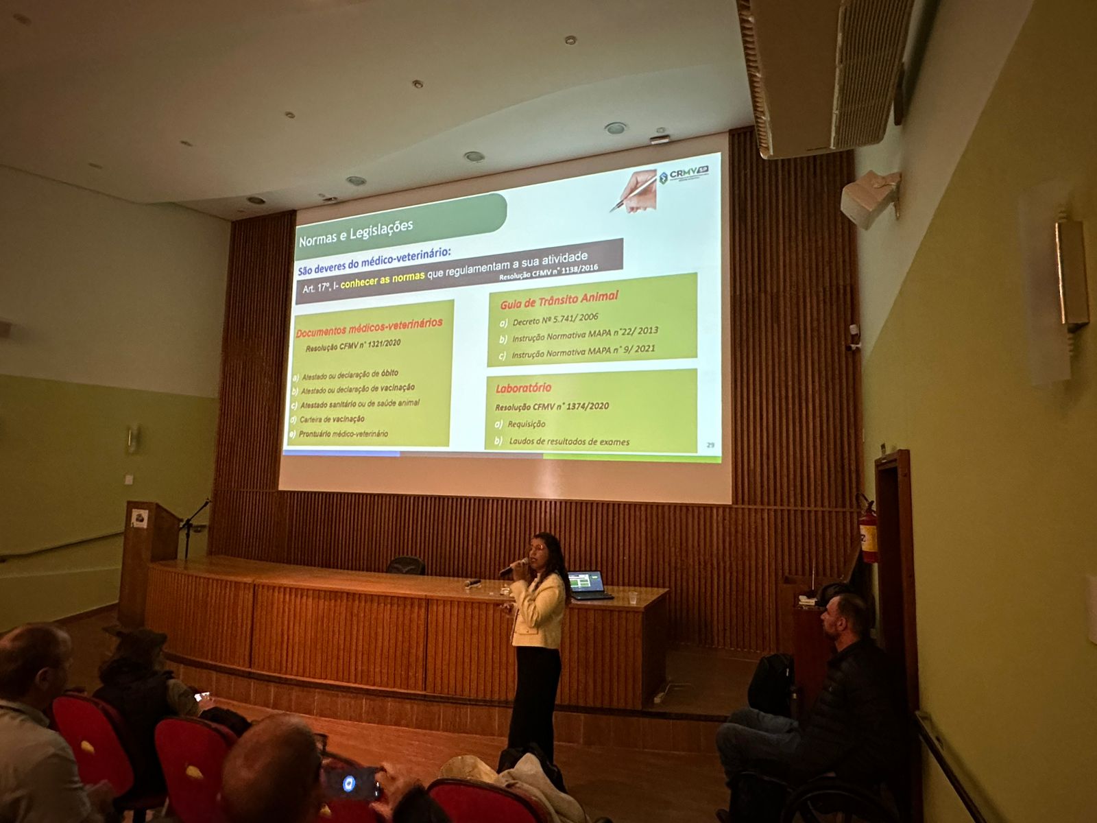 Mulher falando no microfone frente ao telão no palco. Ela veste calça preta e blazer creme com botões dourados. É a dra. Carla Maria Figueiredo.