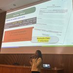 Mulher palestrando frente ao telão. Ela veste calça preta e blazer creme com botões dourados. É a dra. Carla Maria Figueiredo.