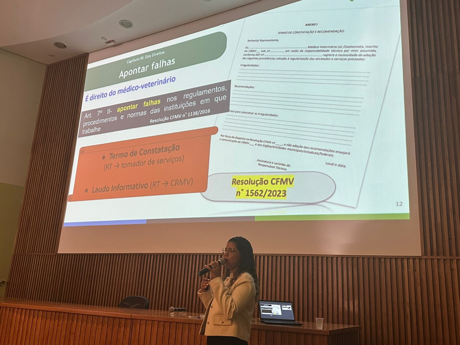 Mulher palestrando frente ao telão. Ela veste calça preta e blazer creme com botões dourados. É a dra. Carla Maria Figueiredo.