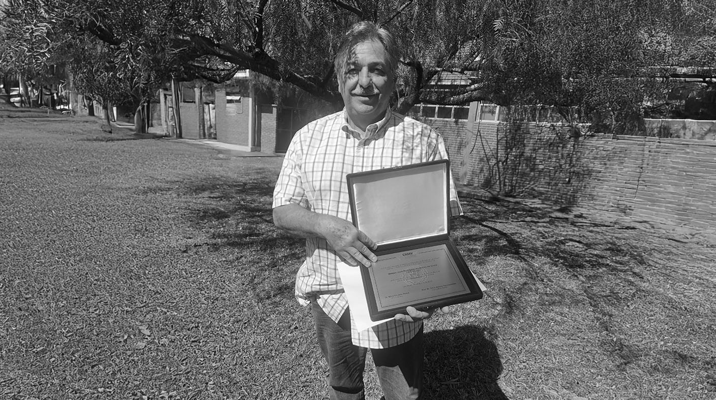 Um homem vestindo camisa de manga curta xadrez, no centro da imagem, segurando uma placa de reconhecimento e, ao fundo, casas e uma árvore fazendo sombra. É o dr. Mateus José Paranhos da Costa.