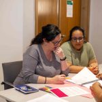 Duas mulheres de óculos e cabelo escuro olhando folha juntas na mesa.
