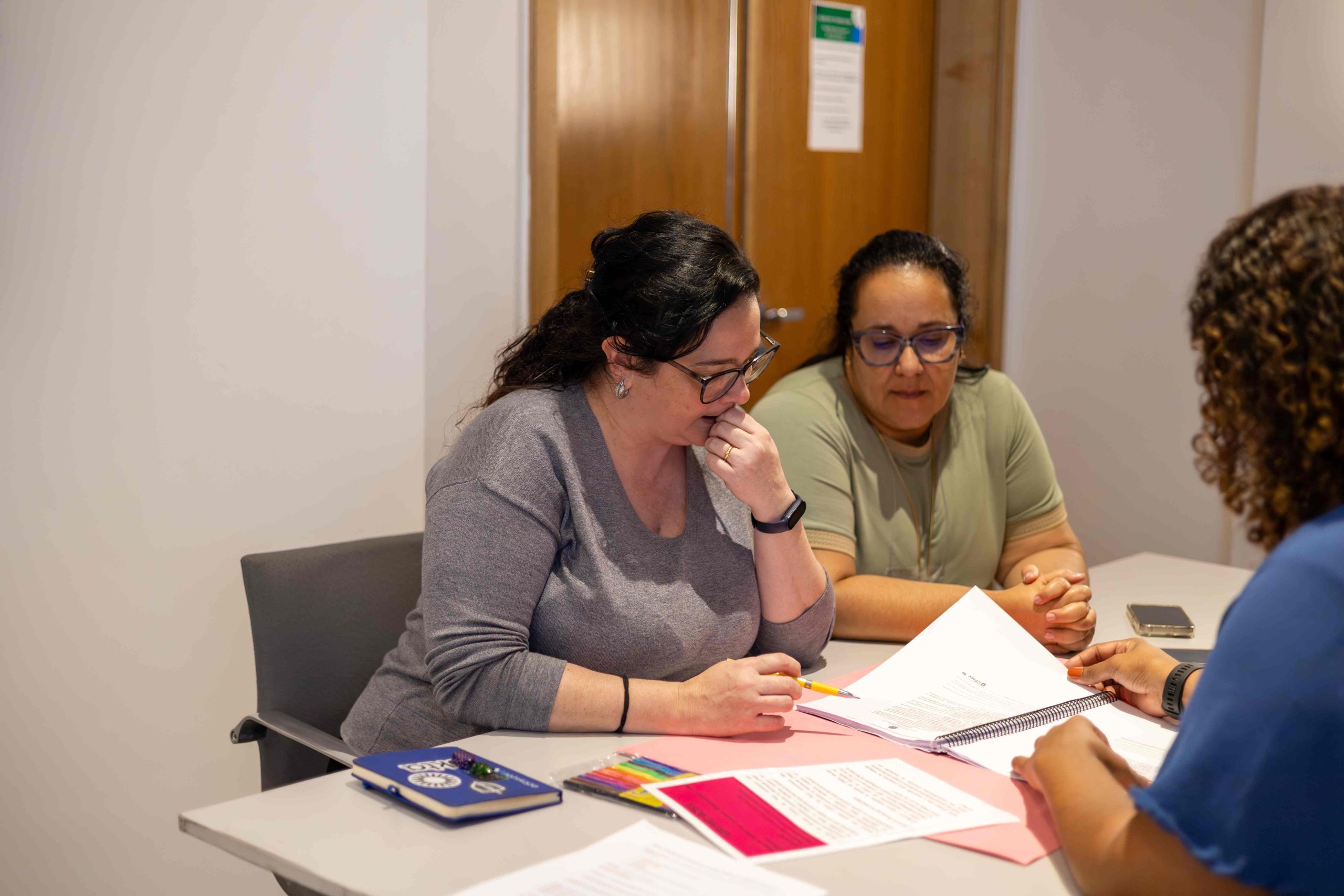 Duas mulheres de óculos e cabelo escuro olhando folha juntas na mesa.