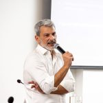 Homem de cabelo grisalho e camiseta branca falando ao microfone, Esse é o Daniel Costa, diretor executivo da Agência BWG.in