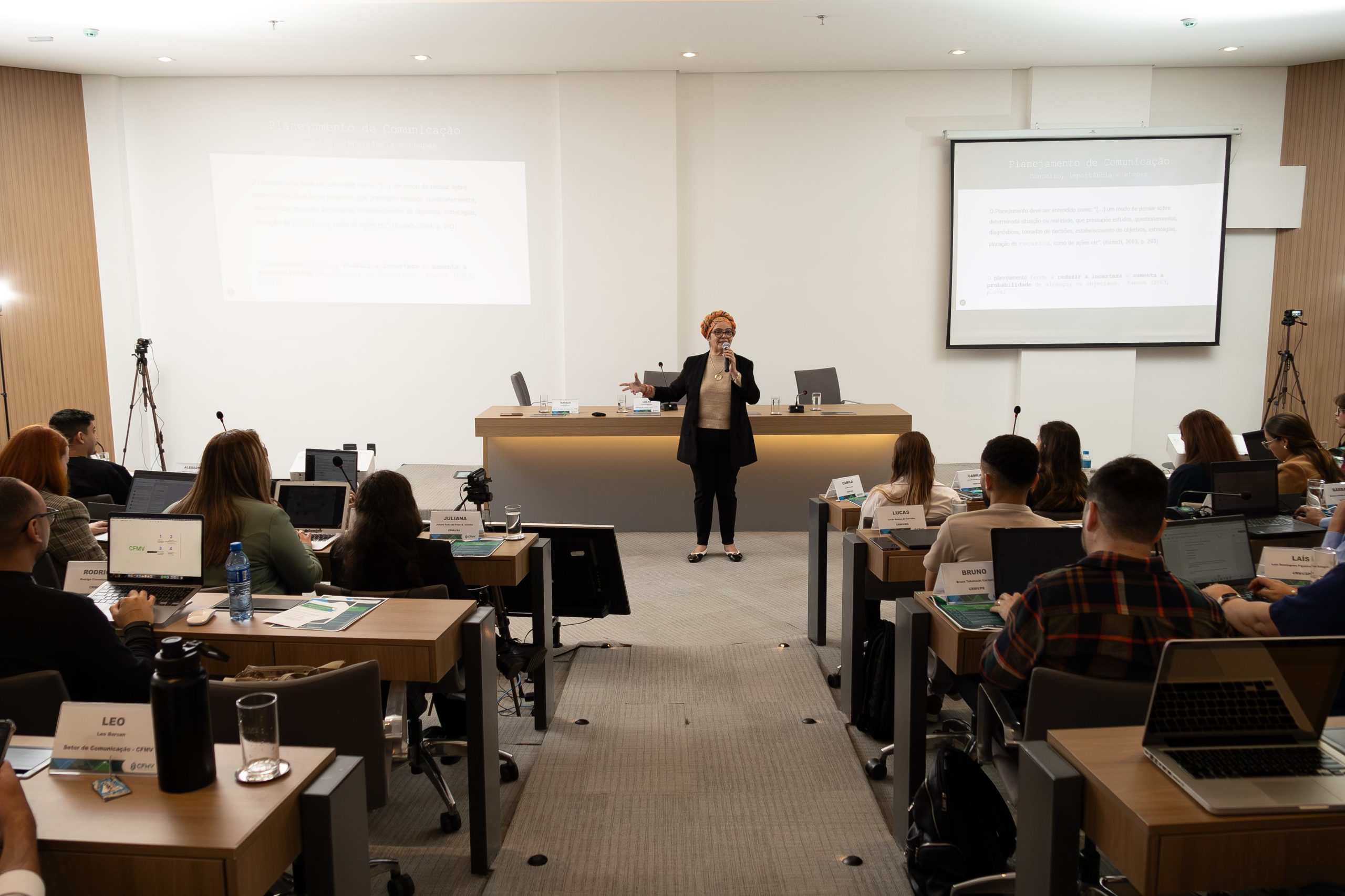 Mulher falando frente ao público sentado.