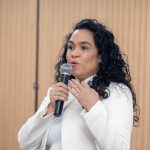 Mulher de cabelo preto e blusa branca falando ao microfone.