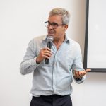 Homem grisalho de óculos, camisa azul falando ao microfone.