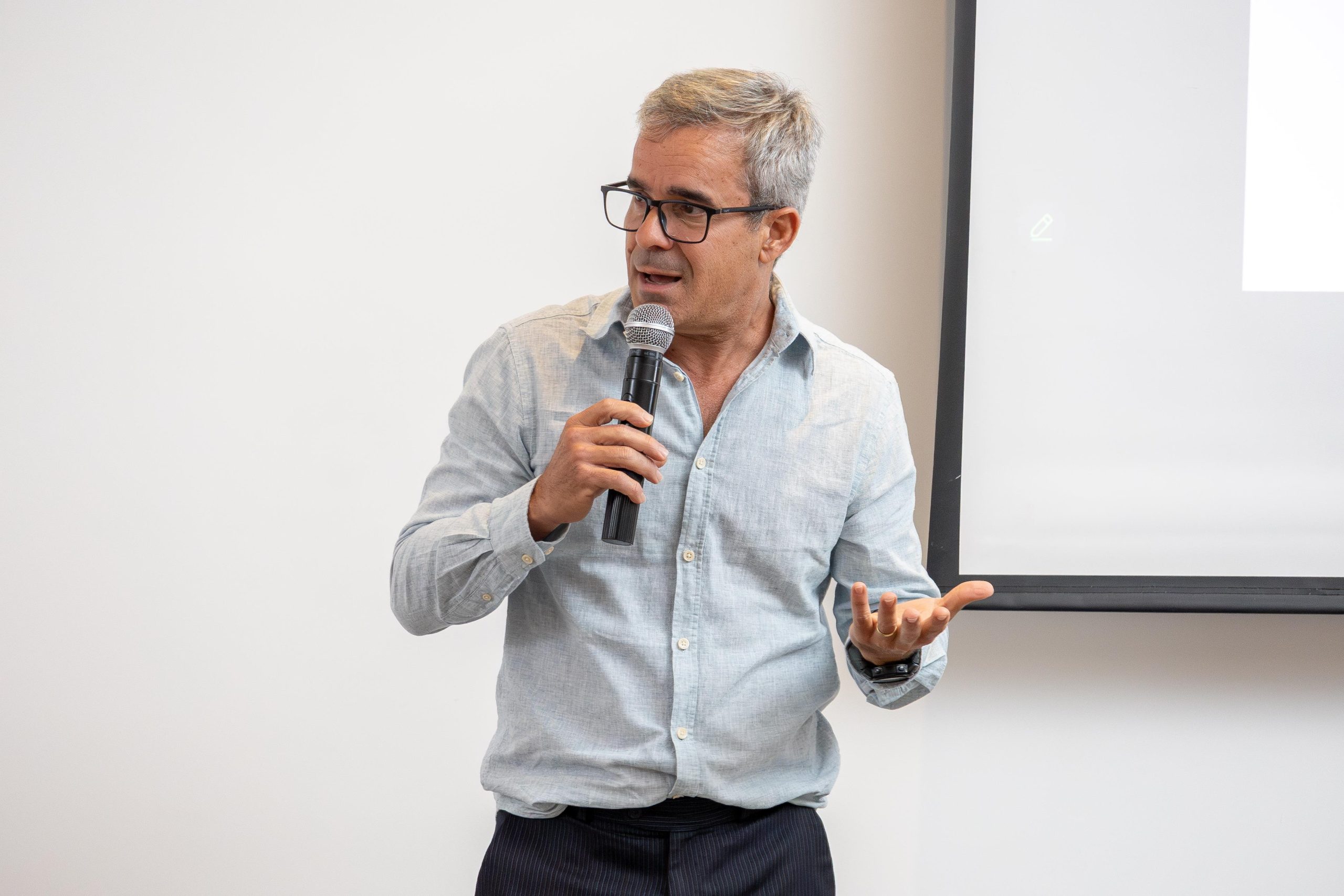 Homem grisalho de óculos, camisa azul falando ao microfone.