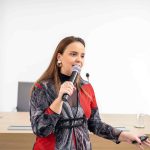 Mulher de cabelos ruivos e blusa cinza com vermelho falando ao microfone.