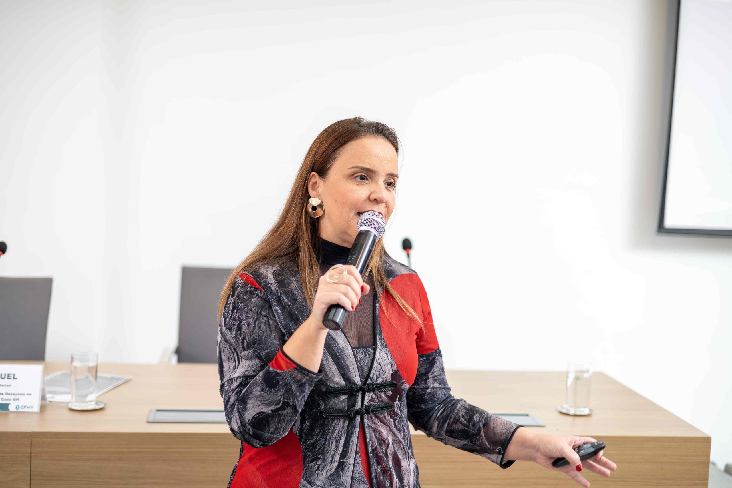 Mulher de cabelos ruivos e blusa cinza com vermelho falando ao microfone.