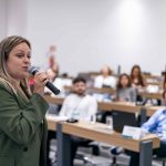 Mulher falando ao microfone para público.