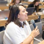 Na foto há uma mulher de cabelo castanho e camiseta branca falando ao microfone.