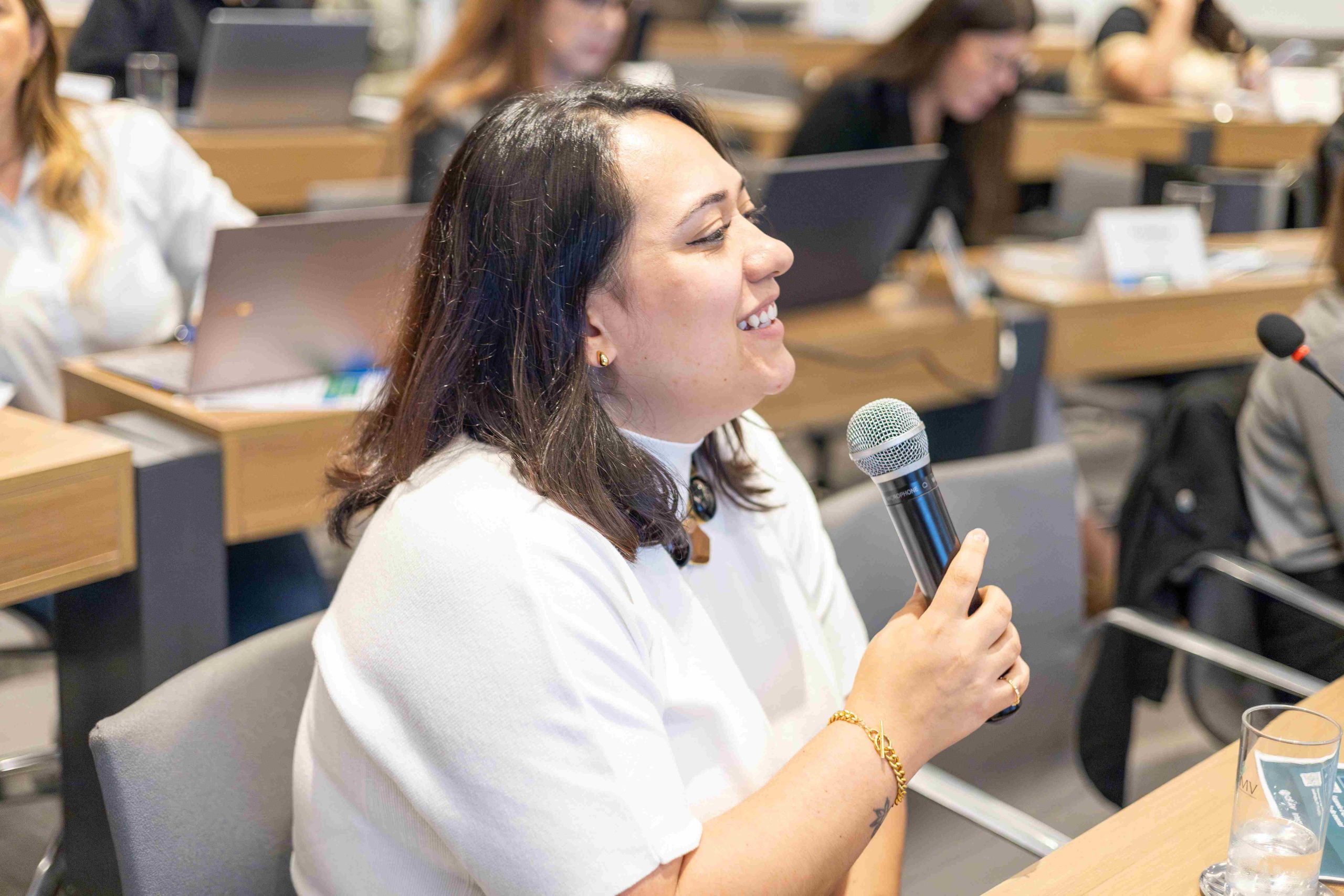 Na foto há uma mulher de cabelo castanho e camiseta branca falando ao microfone.