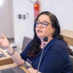 Na foto há uma mulher de cabelo pretos e longo, óculos e camiseta azul. É a dra. Ana Helena Pagotto.