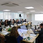 Pessoas reunidas em mesa a frente de um telão.