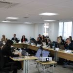 Pessoas reunidas em mesa a frente de um telão.