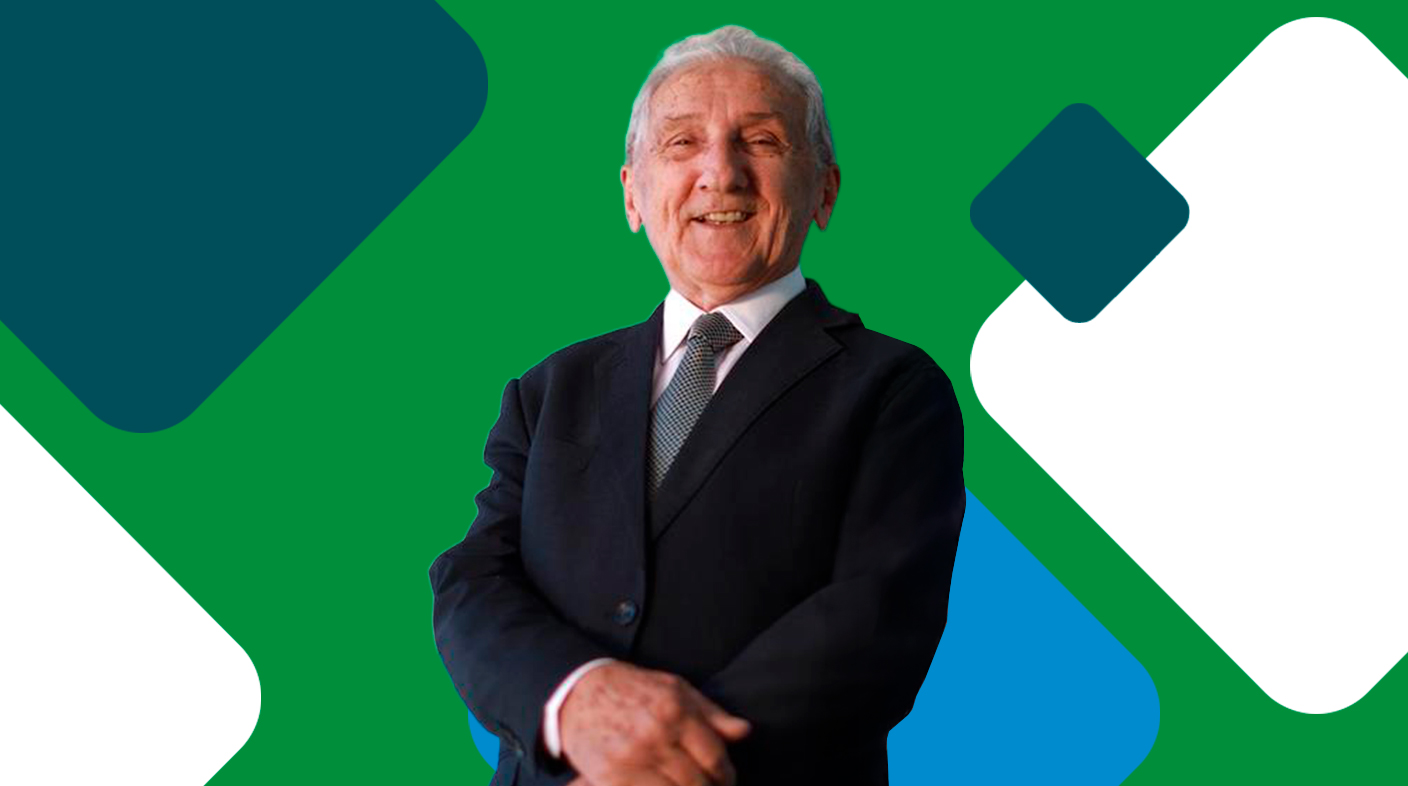 Homem idoso em pé sorridente para foto. Ele veste terno preto com camisa branca e gravata com estampa de listras. Ao fundo da imagem, losangos nas cores institucionais do CFMV: verde, azul, cinza e branco.