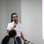 Mulher branca de fios lisos e escuros falando ao microfone na sessão do CFMV. Ela veste calça jeans azul escura e blusa branca de botões no meio com nome da Inataa.